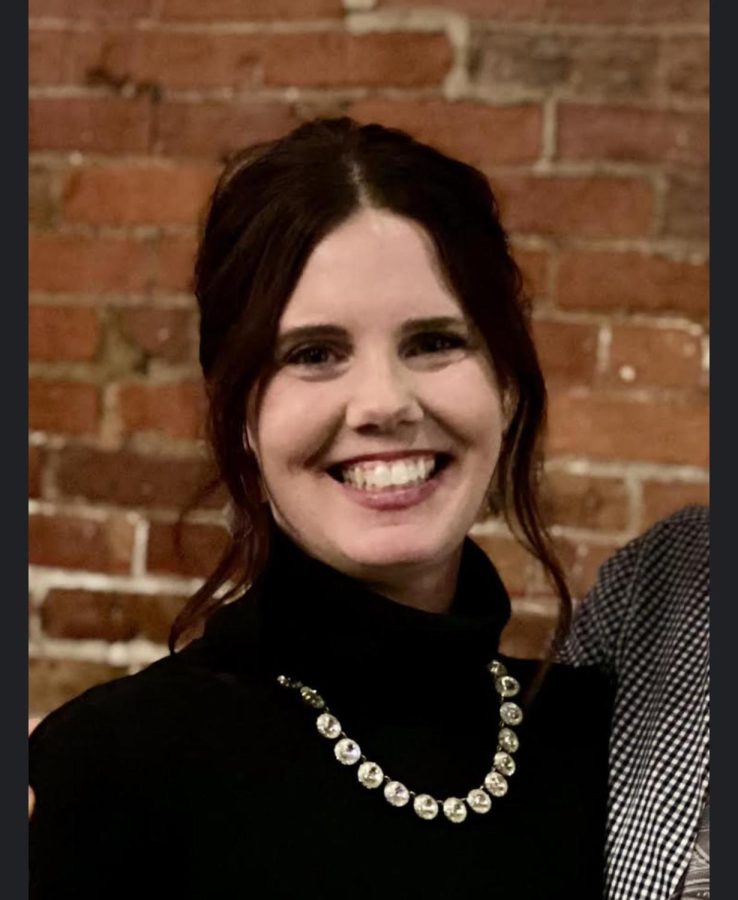 Mrs. Cottingim standing and smiling in front of a brick wall, wearing a black turtleneck with a silver necklace. Her hair is brown and pulled back, leaving only two strands in the front.